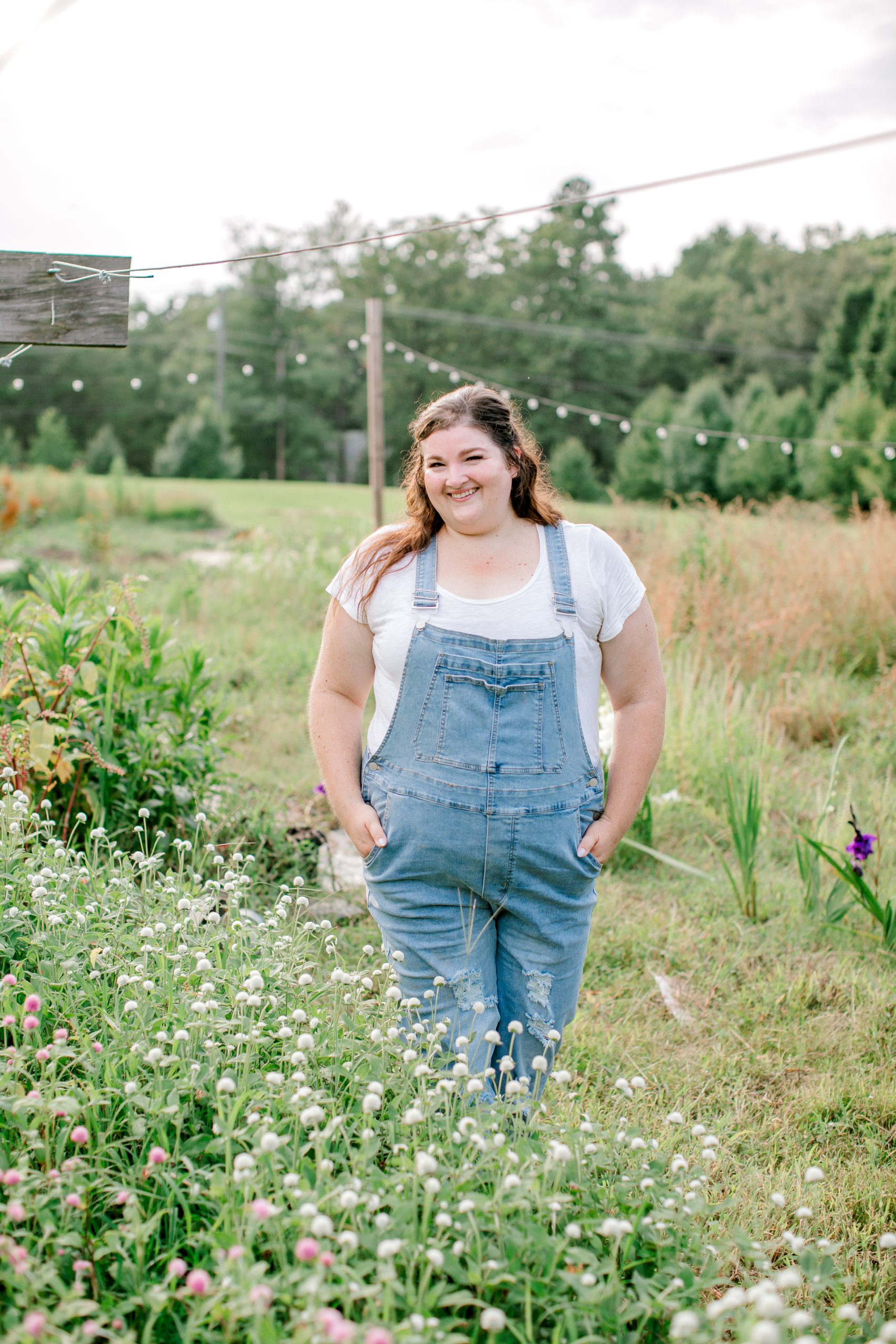 Jamie of Lael Florals, wearing denim overalls, smiles as she stands in her flower garden among rows of white and pink flowers.