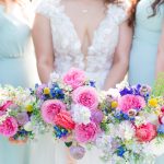 A bride in a lace gown is flanked by two bridesmaids in light blue dresses. They are holding a wide, lush arrangement of pink roses, blue delphiniums, and other colorful wildflowers.