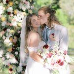 A bride and groom stand in front of a dense floral arch. The groom kisses the bride's cheek, and she smiles while holding a large bouquet that matches the arch's pink, white, and deep red flowers.