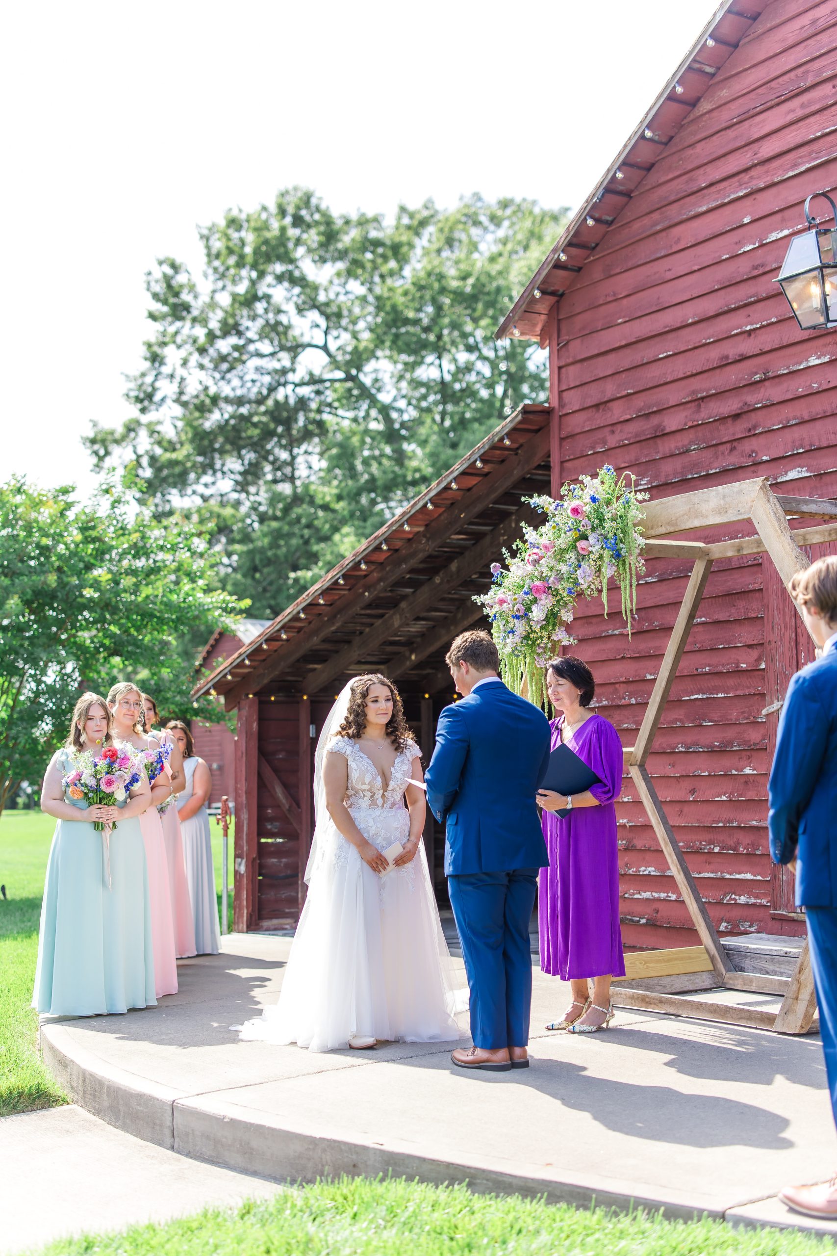 An outdoor wedding ceremony at a red barn featuring a wooden hexagon arch adorned with cascading pastel florals in pink, blue, and green. The bride in a white lace gown and veil stands facing the groom in a blue suit, with bridesmaids in soft pastel dresses holding colorful bouquets lined up behind her.