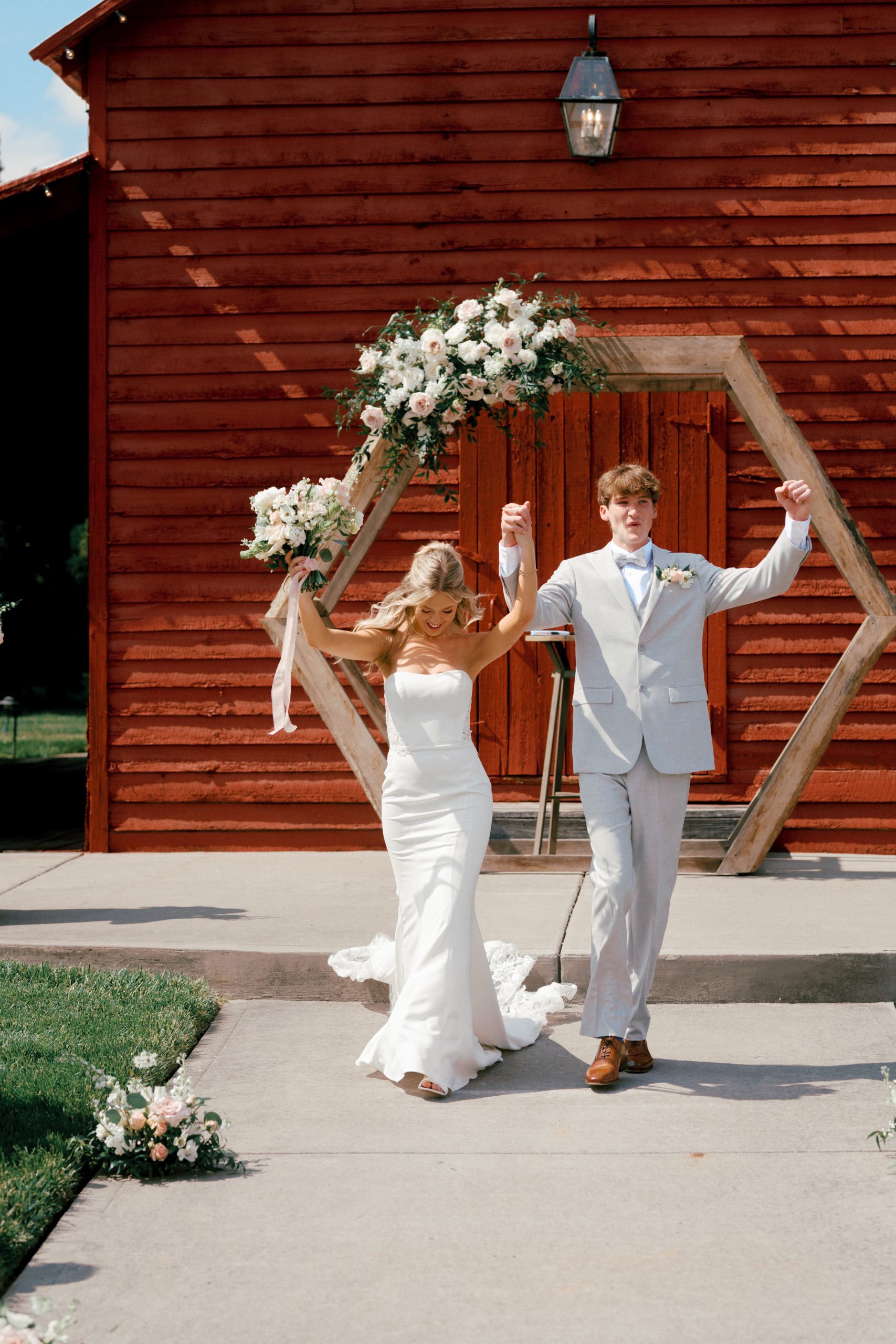 The bride and groom celebrate as they walk down the aisle after their ceremony at The Barns of Kanak. Behind them, a wooden hexagon arch adorned with white and blush florals complements the red barn backdrop. The bride holds her bouquet high, smiling in a fitted white gown, while the groom in a light gray suit raises his arm in joy.