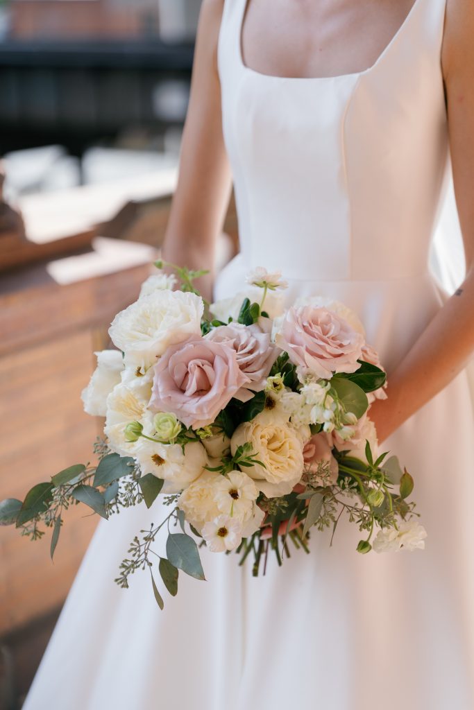 A close-up shot of a bride wearing a modern square-neck wedding gown holding a lush bouquet. The floral arrangement features white garden roses, dusty mauve roses, and white ranunculus mixed with textured greenery.
