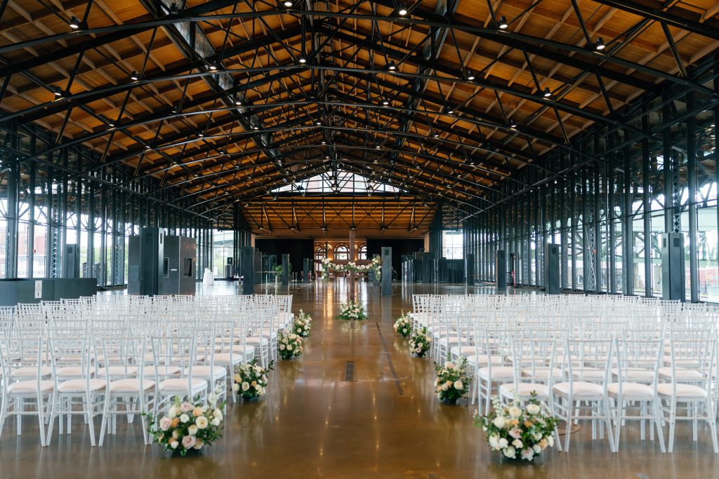 A wide-angle view of an empty wedding ceremony setup inside a massive industrial hall. Rows of white Chiavari chairs flank a polished concrete aisle lined with low floral arrangements. At the far end, a wooden cross decorated with flowers serves as the altar backdrop beneath a high ceiling with exposed black metal trusses and wood paneling.