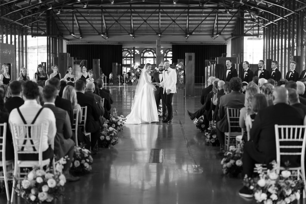 A wide-angle, black-and-white photograph of a wedding ceremony inside a large industrial venue. The bride and groom stand at the altar in the distance, smiling at one another; the groom wears a white tuxedo jacket. Guests are seated in rows on either side of a polished aisle lined with low floral arrangements. The high ceiling features exposed steel trusses.