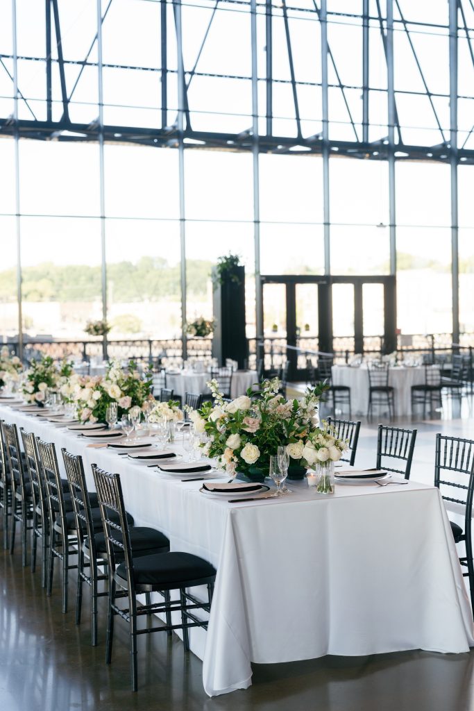 A long, rectangular head table at a wedding reception set with white linens and black Chiavari chairs. The table is adorned with low, lush floral centerpieces featuring white and dusty mauve roses mixed with greenery. The background features a massive wall of floor-to-ceiling industrial windows with black steel framing, letting in bright natural light.